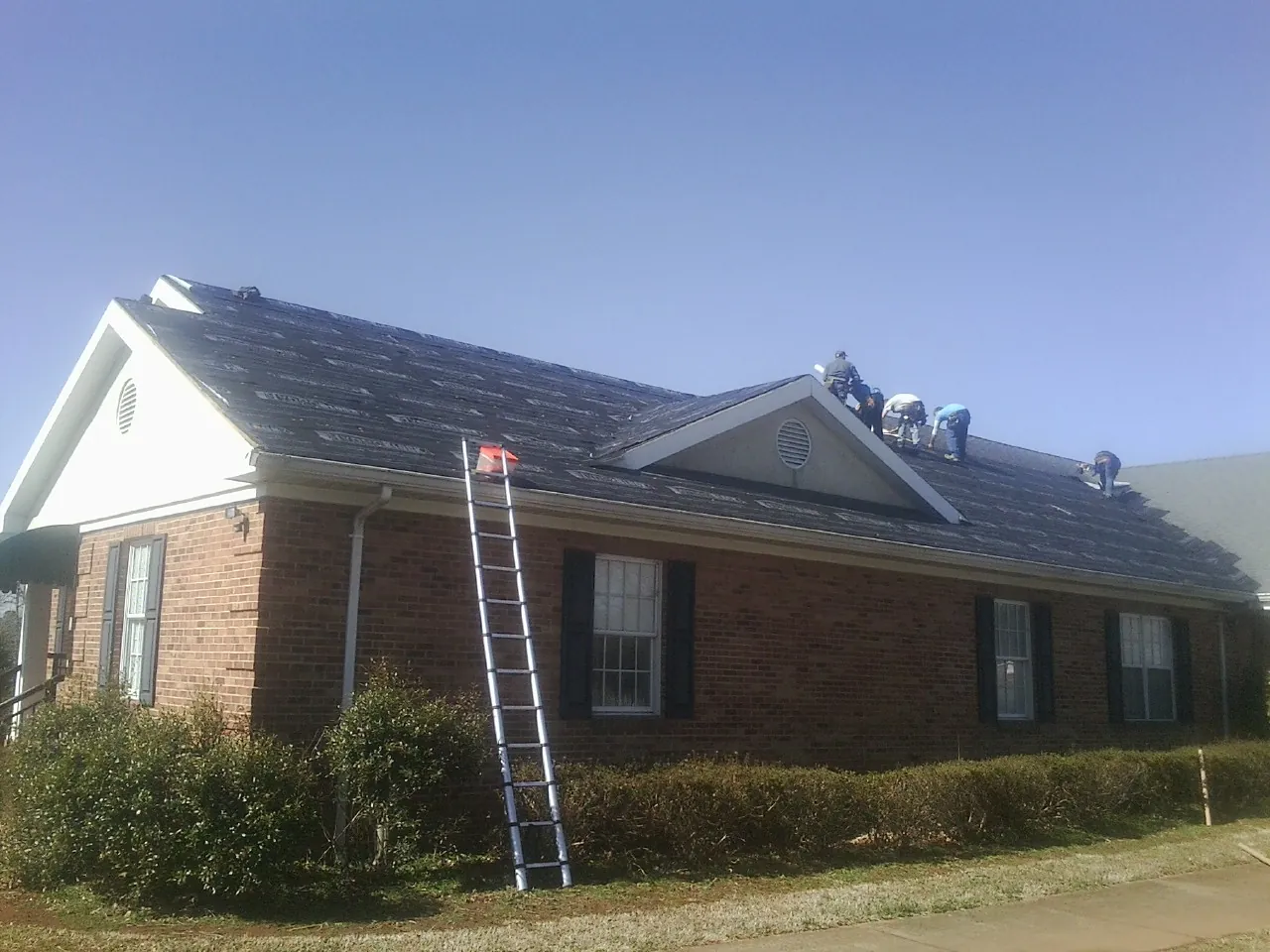 Crew tearing shingles off a brick ranch roof with a ladder set — Emergency Roof Repair work by our crew in Brice