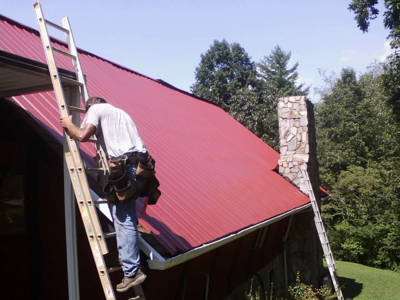 Roofer on a ladder beside a red metal roof and stone chimney — Soffit & Fascia Repair by Roofing Brothers in Brice